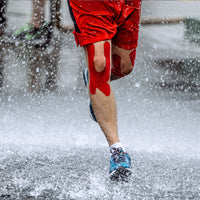 Runner wearing red TapeGeeks kinesiology tape on knees in rain, showing strong, water-resistant muscle and joint support.