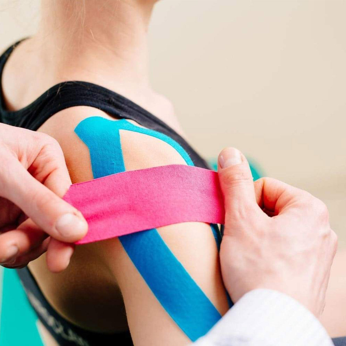 Close-up of a therapist applying pink kinesiology tape over blue tape for shoulder alignment and muscle support.

