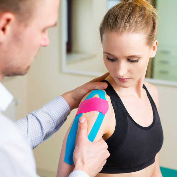 A physical therapist applying pink and blue TapeGeeks kinesiology tape to a woman’s shoulder for muscle support and pain relief.